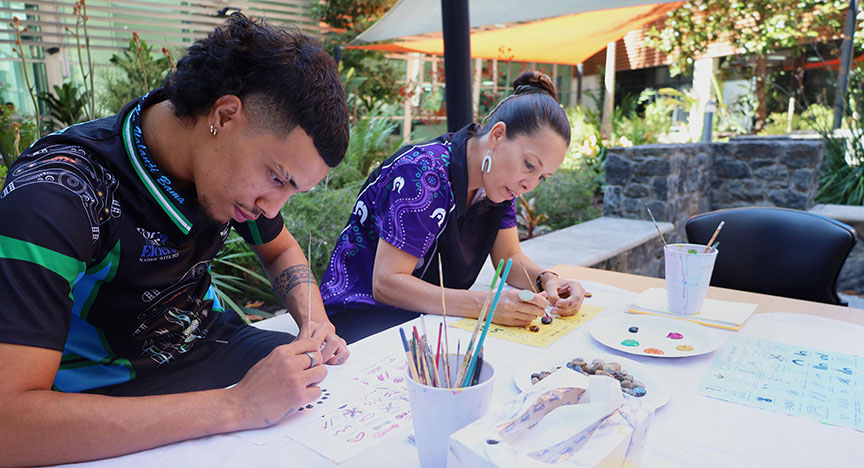 Two First Nations individuals engage in an outdoor art activity, painting and crafting at a table with art supplies.