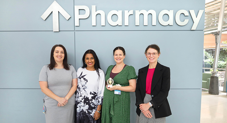 Four people stand in front of a wall with a Pharmacy sign. One person holds a trophy.