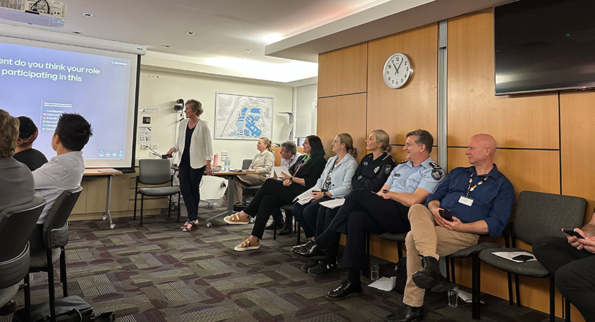 A group of people in a meeting room, with a presenter standing near a screen displaying a question. Some attendees are in uniform.