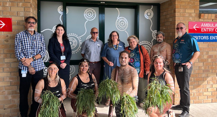 A group of people, including First Nations individuals holding greenery, stand in front of a building with decorated glass doors.