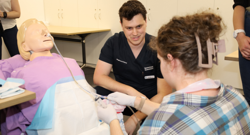 A healthcare worker demonstrates a medical procedure on a mannequin while others observe in a clinical setting.