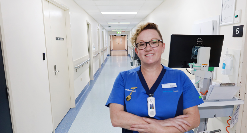 A nurse in blue scrubs stands in a hospital corridor with arms crossed, next to medical equipment.