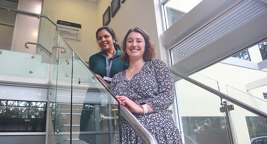 Two people standing on a staircase inside a building with a Metro South Health sign in the background.