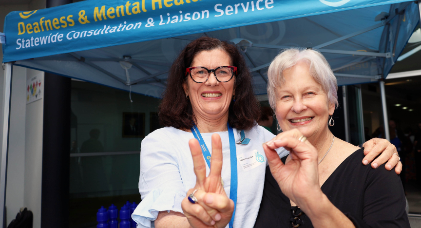 Two people stand in front of a tent labeled Deafness & Mental Health Statewide Consultation & Liaison Service, making hand signs.