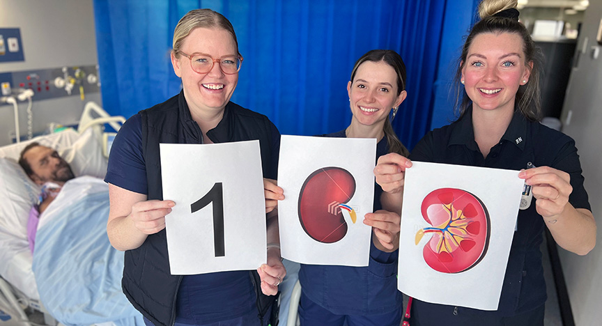 Three healthcare workers hold signs showing 100 with kidney illustrations, celebrating the 100th kidney transplant for 2023.