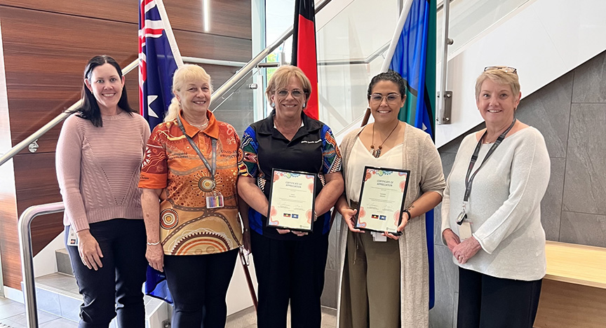 Five people standing indoors, two holding certificates. Australian and First Nations flags are in the background.