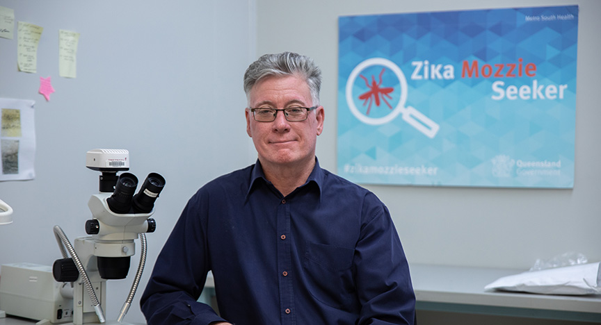 A person sits in a lab with a microscope. A sign behind reads Zika Mozzie Seeker from Metro South Health, Queensland.