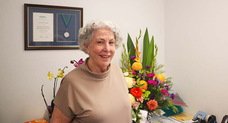 An elderly person stands in an office with a framed certificate and medal on the wall, and a colorful bouquet of flowers on the desk.