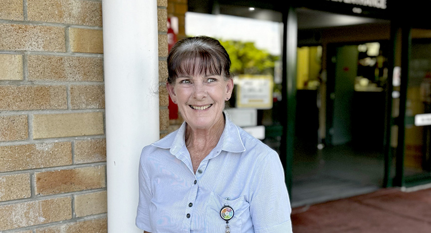 A person in a light blue uniform stands outside a building with a sign reading Emergency.