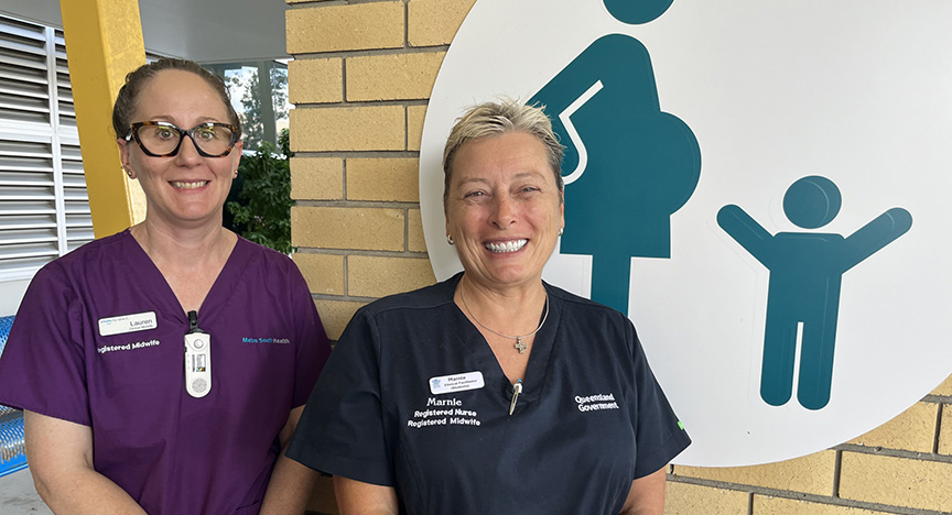 Two registered midwives in uniforms stand in front of a healthcare facility sign depicting a pregnant person and a child.