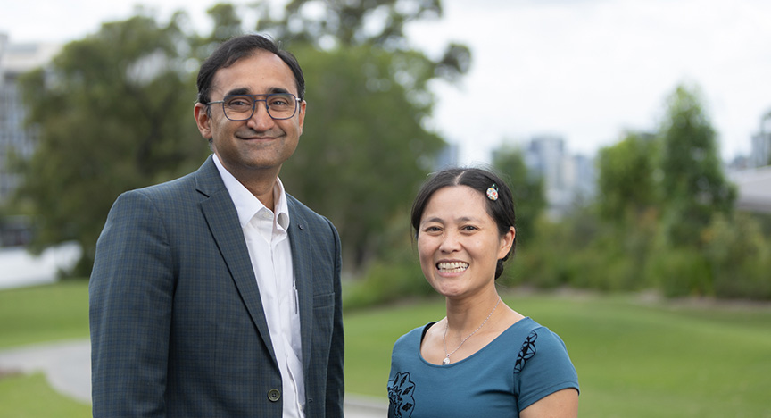 Two people standing outdoors in a park, with trees and buildings in the background. One person is wearing a suit, and the other is in a blue top.