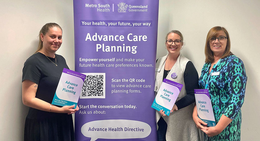 Three people holding Advance Care Planning brochures stand beside a banner promoting advance care planning by Metro South Health and Queensland Government.