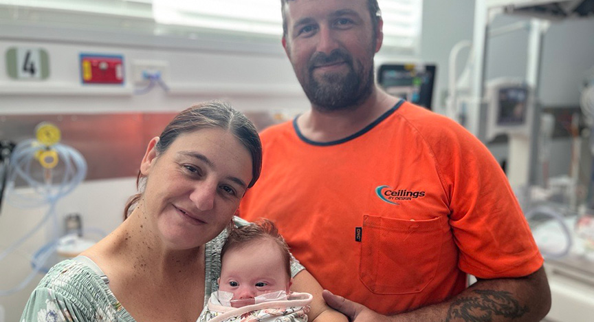 Two adults in a hospital room, one holding a baby. Medical equipment is visible in the background.