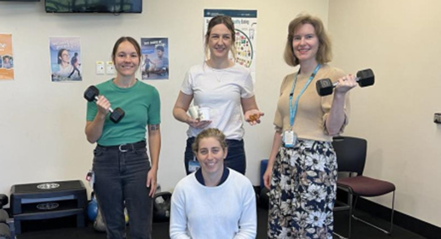 Four people in a room with exercise equipment, two holding dumbbells, one holding a pill bottle, and one kneeling. Posters are on the wall behind them.