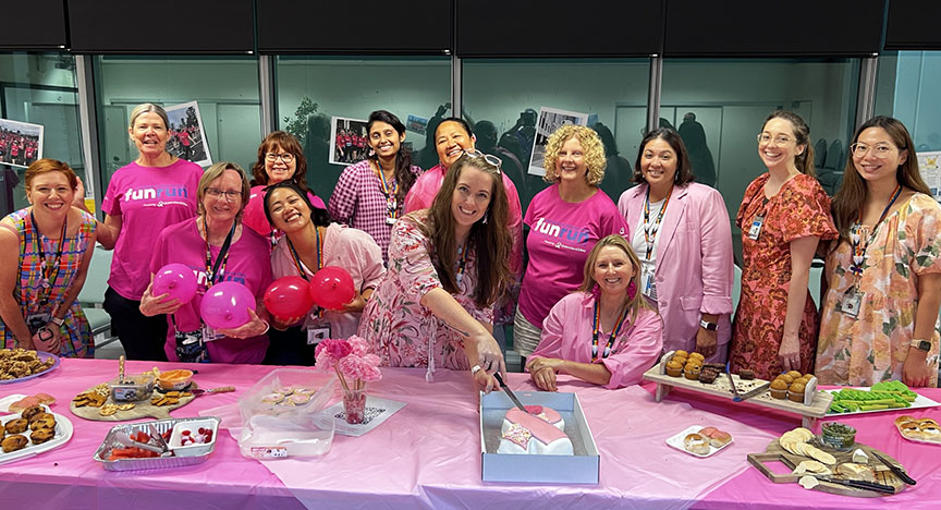 A group of people in pink attire gather around a table with food, celebrating a breast cancer awareness event.