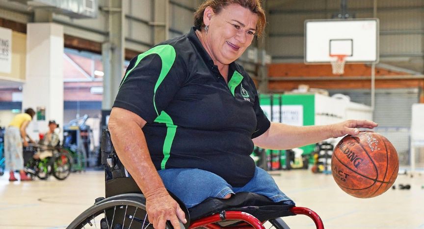 A person in a wheelchair holding a basketball in an indoor sports facility. Other people in wheelchairs are visible in the background.