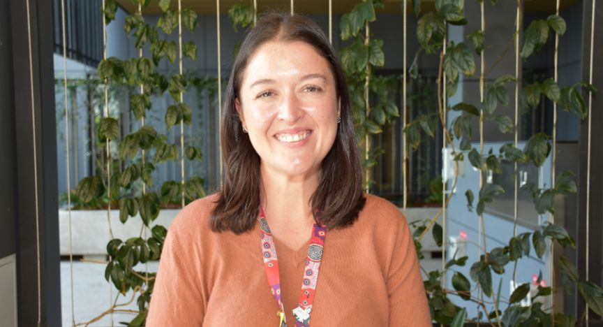 A person wearing a brown sweater and a colorful lanyard stands in front of a green, leafy background.