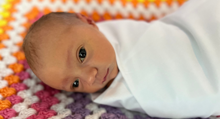 A baby wrapped in a white blanket lies on a colorful crocheted blanket with orange, pink, purple, and white patterns.