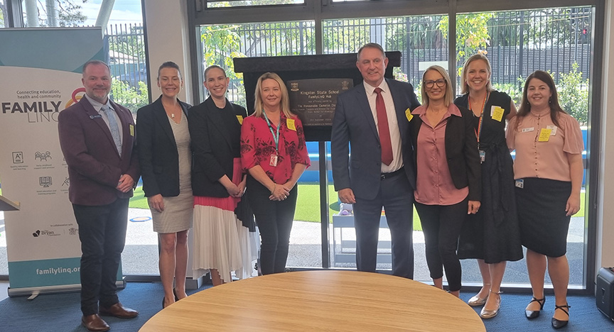 Eight staff members from Metro South Health and the Department of Education smiling in front of a plaque at the official opening of FamilyLinQ at Kingston State School, wearing corporate clothing.