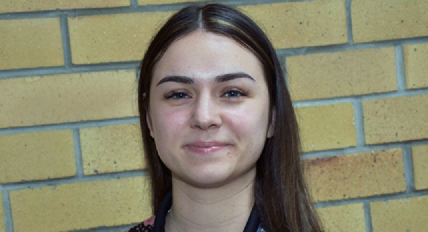 A person with long hair stands in front of a yellow brick wall.