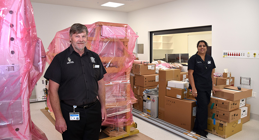 Two people in black uniforms stand in a room filled with large medical equipment covered in pink plastic and numerous cardboard boxes.