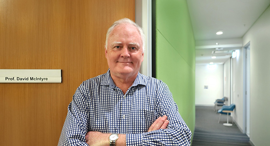 A person stands with arms crossed in front of a door labeled Prof. David McIntyre in a hallway with green and white walls.