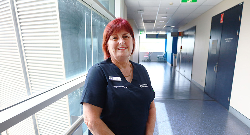 A nurse stands in a hospital corridor, wearing a uniform with Registered Nurse and Queensland Government badges.