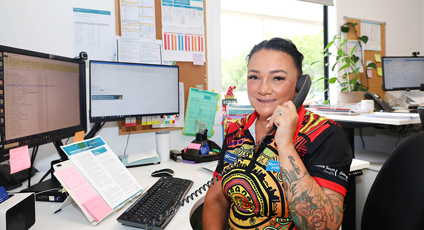 A person in a colorful shirt is on the phone at a desk with multiple computer monitors and office supplies.