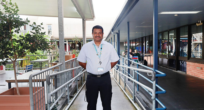 A person stands in a covered outdoor walkway, wearing a white shirt and a lanyard with an ID badge. The walkway has railings and potted plants.