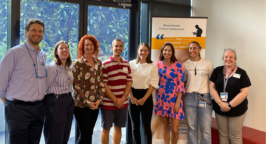 A group of eight people standing indoors in front of a banner for the Mental Health Clinical Collaborative.