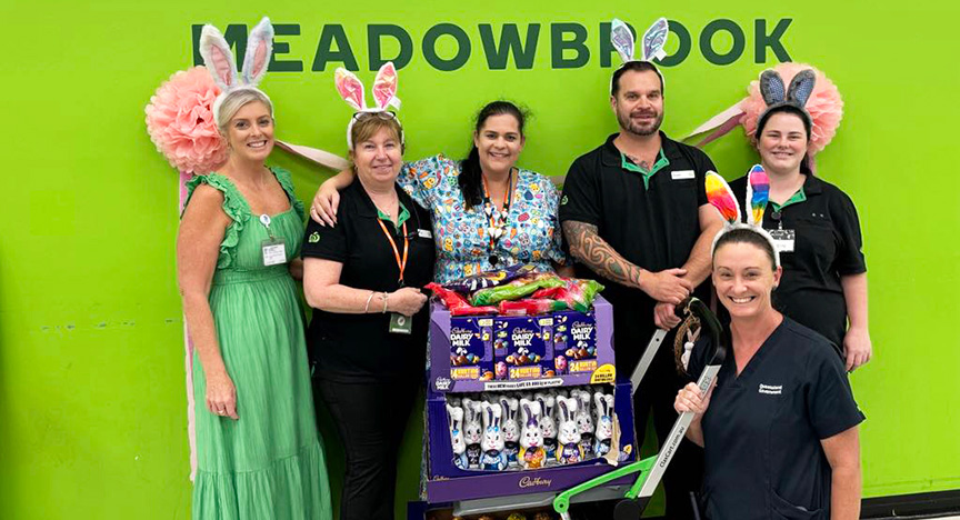 A group of people in festive attire stand around a display of Easter chocolates in front of a green wall with MEADOWBROOK written on it.