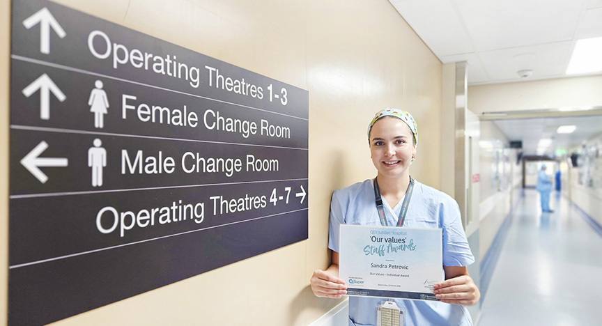 A nurse in scrubs holds a certificate in a hospital hallway near a sign indicating directions to operating theatres and change rooms.