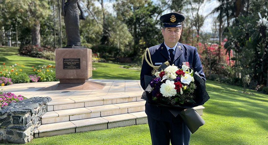 A person in military uniform holds a floral wreath in front of a memorial statue in a park with flowers and trees.