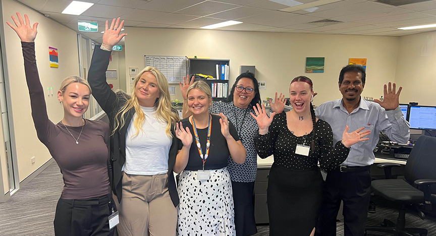 A group of six people in an office, smiling and raising their hands, celebrating.