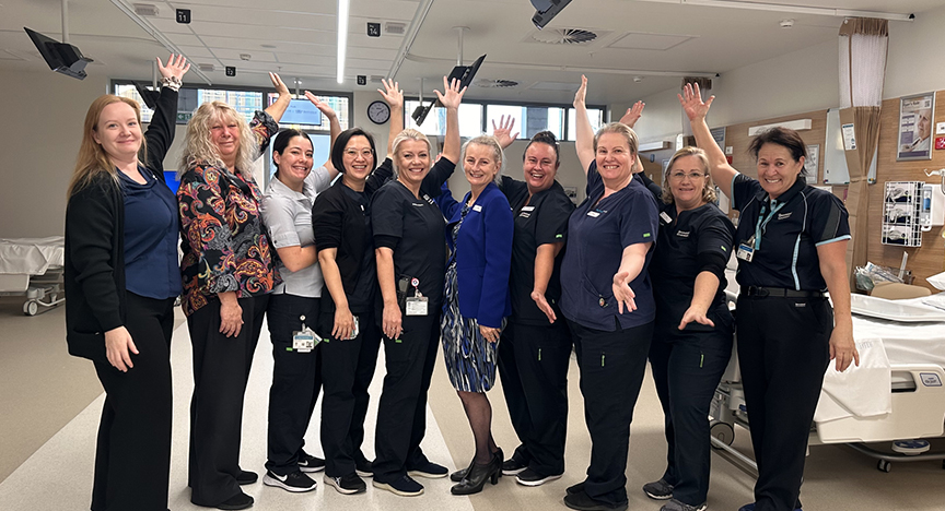 A group of healthcare workers in a hospital room, standing in a semi-circle with raised hands, smiling and celebrating.