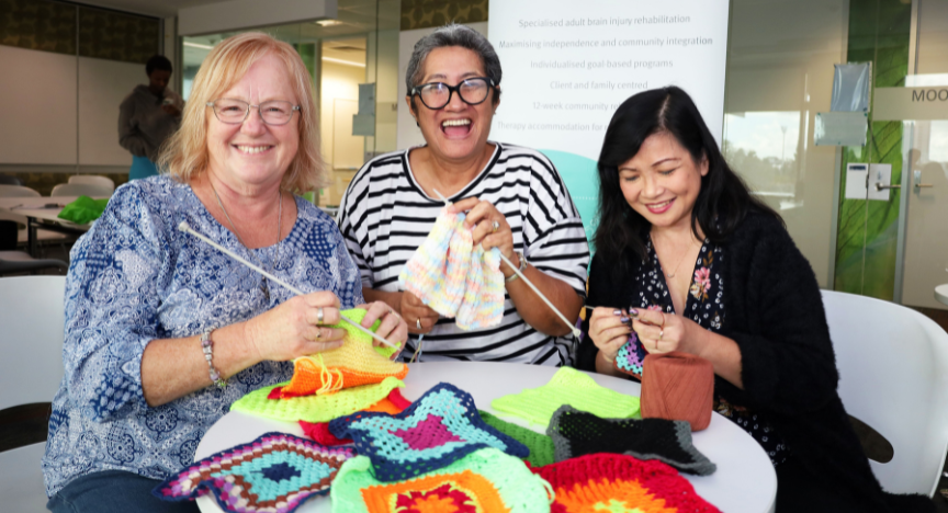 Three people knitting colorful squares at a table, with a banner about brain injury rehabilitation in the background.