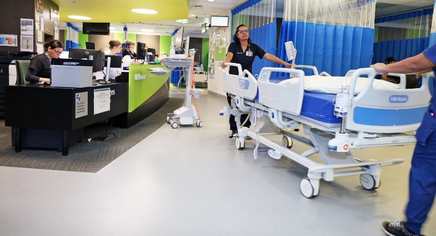 Hospital staff at a nursing station, with two healthcare workers moving an empty hospital bed in a brightly lit corridor.