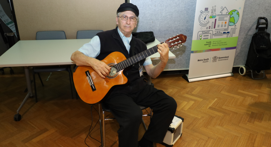 A person playing a guitar sits in a room with a table and chairs. A banner for Metro South Health and Queensland Government is in the background.