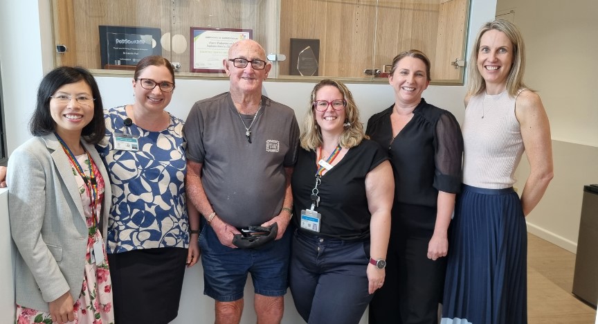 five staff members and one patient standing in front of a cabinet, wearing a mix of casual and smart casual attire. 