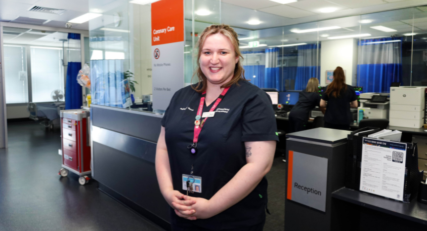 Megan Seymour, Cardiology NUM, stands in her hospital uniform in front of a desk in the Cardiac Care Unit