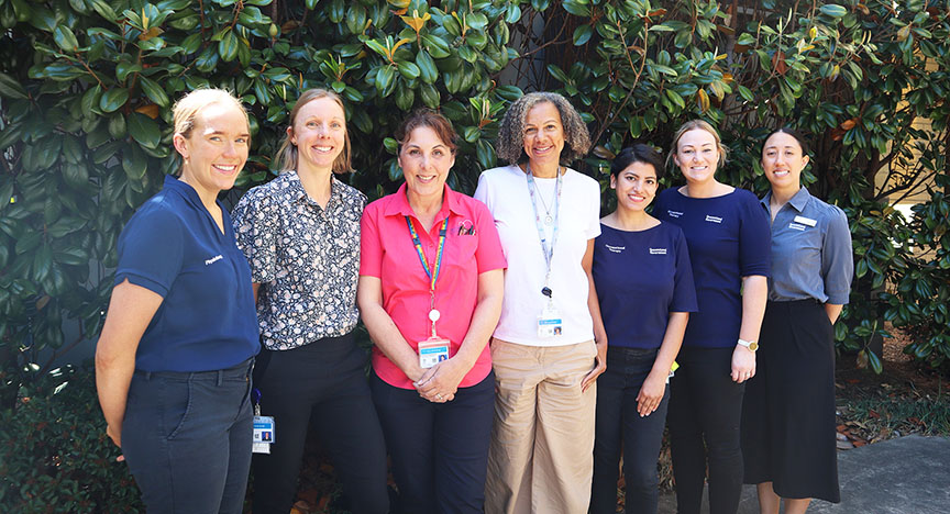 A group of seven healthcare workers stands in front of a leafy background, wearing uniforms and name badges.