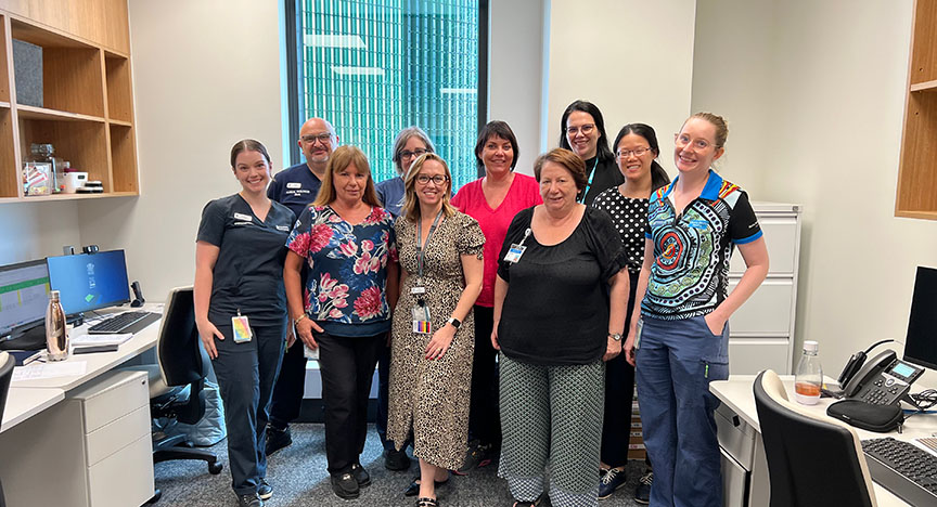 A group of healthcare professionals standing in an office, posing for a photo. The office has desks, computers, and shelves.