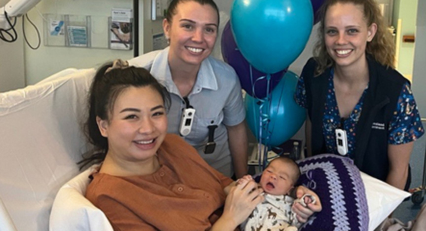 A person in a hospital bed holding a baby, with two healthcare workers and balloons in the background.