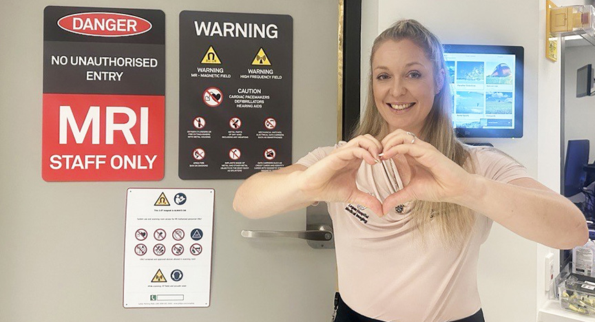 A person stands in front of an MRI room door with warning signs, making a heart shape with their hands.