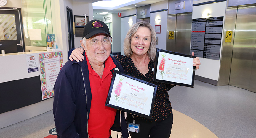 Two people holding framed Metro South Health Volunteer Awards certificates in a hospital lobby.