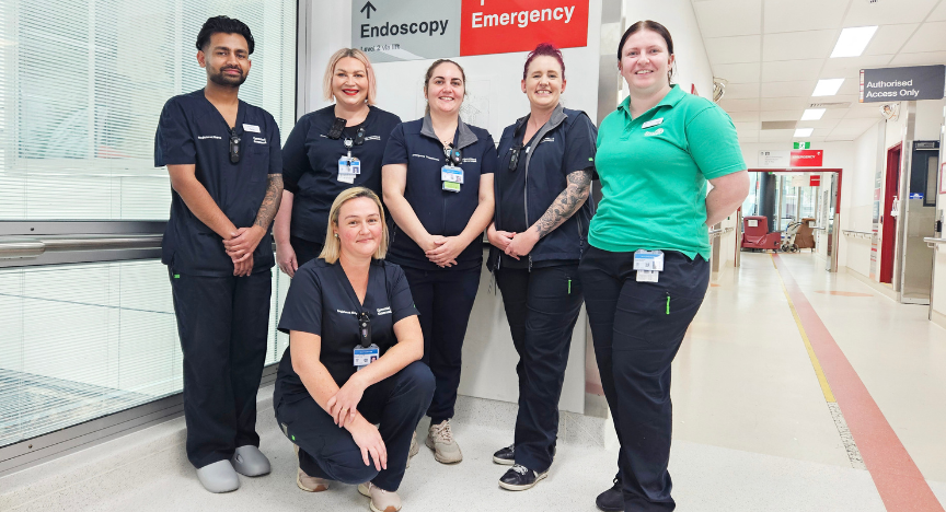 A group of healthcare workers in uniforms stands in a hospital hallway near signs for Endoscopy and Emergency.