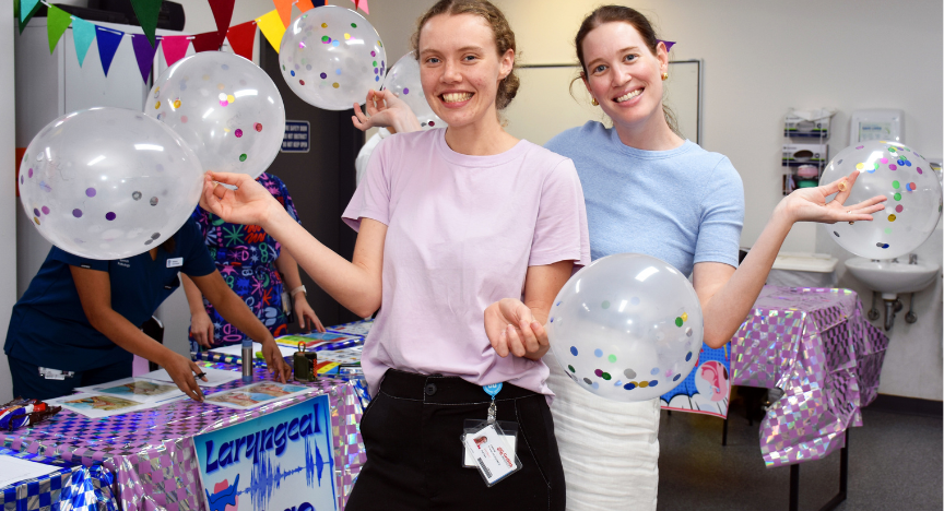 Two people holding balloons at a decorated table with colorful flags and a sign reading Laryngeal.