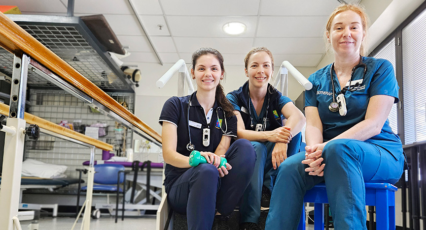 Three healthcare workers in uniforms sit on a staircase in a medical facility, with physiotherapy equipment visible in the background.