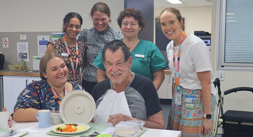 A group of healthcare workers and a patient in a hospital setting, with the patient seated at a table with a meal.