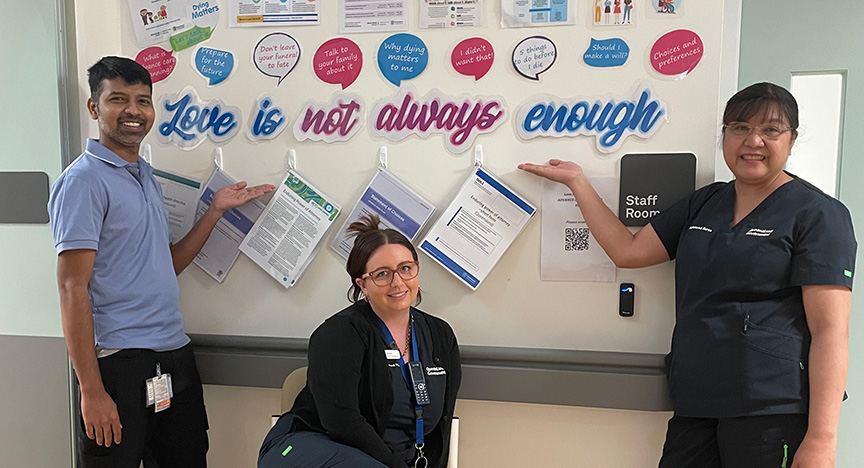 Three healthcare workers in a hospital pose in front of a bulletin board with the message Love is not always enough and various informational posters.